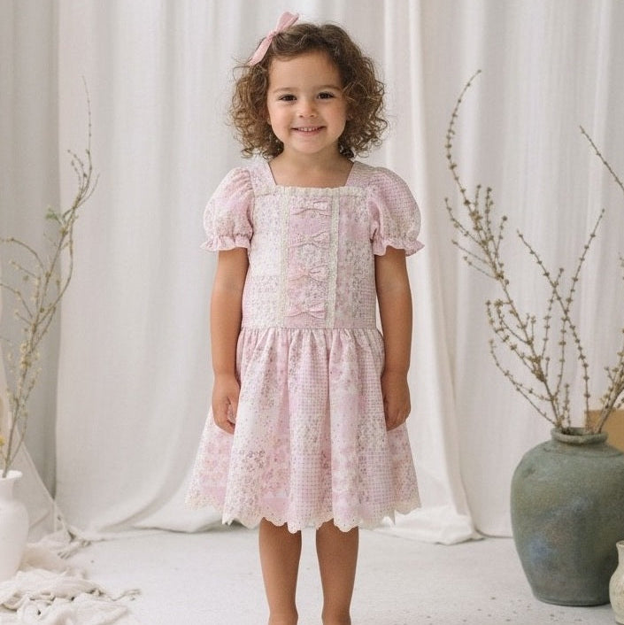 A young girl with curly brown hair wearing a light pink lace dress with puff sleeves and a bow in her hair, standing in front of a white curtain backdrop.