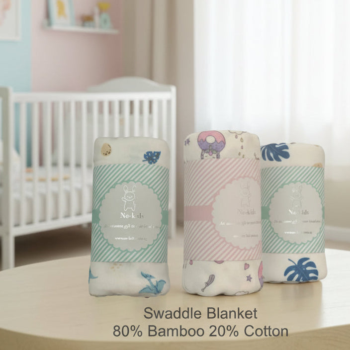 Three rolled swaddle blankets with printed labels are displayed on a light wooden surface in a softly lit indoor setting. The blankets are white with pastel prints: one mint green with blue leaves, one pink with pastel donut and cloud motifs, and one mint green with blue leaves.